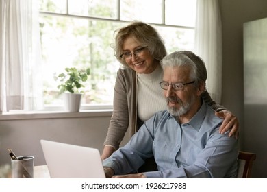 Happy Mature OAP Couple Study Internet App On Laptop, Grandparents Use Computer At Home For Online Shopping. Smiling Middle Aged Pensioner Wife Hugs Senior Husband During Video Call To Relations