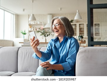 Happy Mature Old 60s Woman Holding Smartphone Using Mobile Phone App For Video Call, Laughing While Watching Funny Video, Feeling Excited Winning Online Lottery Bid On Cellphone Sits On Couch At Home.