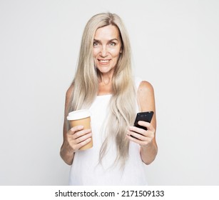 Happy Mature Old 60s Woman With Long Hair Holding Smartphone And Takeaway Coffee Over Grey Background