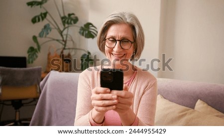Happy mature old 60s woman, older middle aged female holding smartphone using mobile app, texting message while chatting with friends with cell phone on couch at home. Happy laugh, having fun.