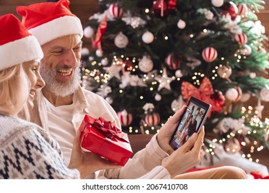 Happy Mature Old Grandparents Couple In Santa Hats Greeting Family Presenting Christmas Gift On Remote Video Call Using Digital Tablet, Having New Year Online Virtual Party Meeting Videocall At Home.