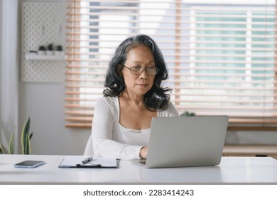 Happy mature old woman remote working from home distance office on laptop taking notes. Smiling 60s middle aged business lady using computer watching webinar and writing in notebook - Powered by Shutterstock