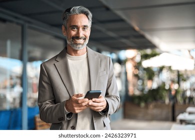 Happy mature older business man executive using mobile cell phone outdoors. Middle aged businessman manager or entrepreneur holding smartphone looking at camera standing outside office. Portrait. - Powered by Shutterstock