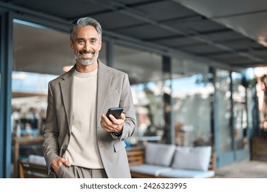 Happy mature older business man entrepreneur wearing suit using mobile cell phone outdoors. Middle aged businessman executive holding smartphone looking at camera standing outside office. Portrait. - Powered by Shutterstock