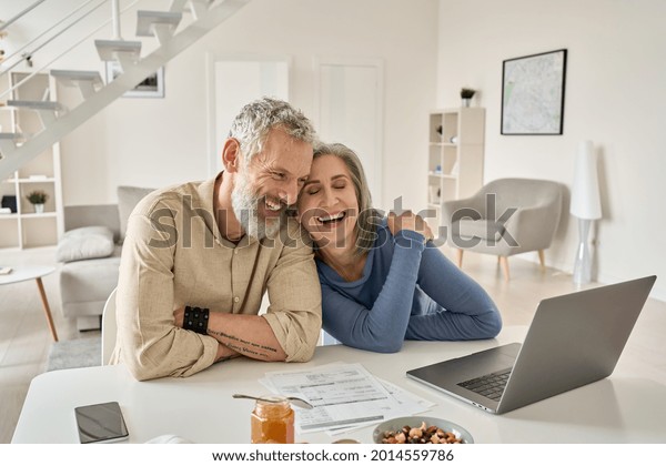 Happy mature older family couple laughing,\
bonding sitting at home table with laptop. Smiling middle aged\
senior 50s husband and wife having fun satisfied with buying\
insurance, paying bills\
online.
