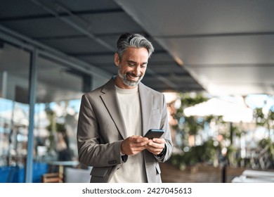 Happy mature older male entrepreneur standing outdoors looking at smartphone. Smiling middle aged business man, rich professional businessman holding cellphone using mobile cell phone digital tech.