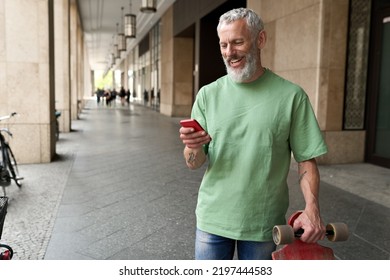 Happy mature older middle aged bearded man using mobile cell phone standing outside. Smiling old adult male hipster with skateboard holding cellphone texting message on smartphone digital technology.