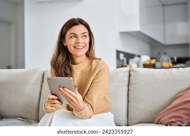 Happy mature older woman using digital tablet sitting on couch at home. Smiling middle aged 45 years old woman looking away at copy space holding tab computer relaxing on sofa in living room. - Powered by Shutterstock