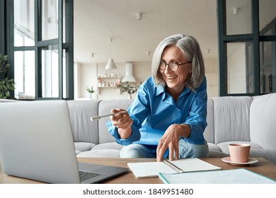 Happy Mature Older Woman Video Calling On Laptop Working From Home. Smiling 60s Middle Aged Businesswoman Talking By Conference Online Virtual Chat Using Computer At Home Office Sitting On Couch.