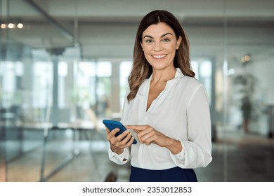 Happy mature professional business woman using cell phone at office, portrait. Smiling mid age 45 years old businesswoman executive standing at work lobby holding smartphone looking at camera. - Powered by Shutterstock