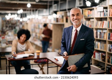 Happy Mature Professor Holding Digital Tablet While Teaching Students At University Library And Looking At Camera.