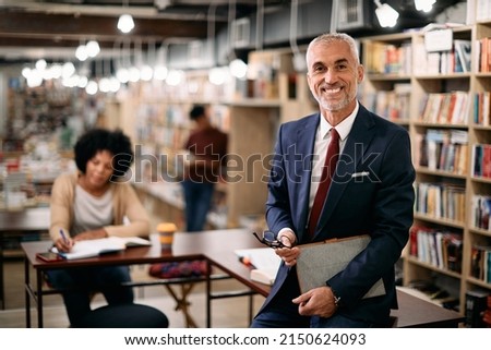 Happy mature professor holding digital tablet while teaching students at university library and looking at camera.