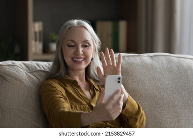 Happy Mature Retired 50s Grandma Woman Waving Hand Hello At Smartphone Frontal Camera, Talking To Family On Video Conference Call, Smiling, Laughing, Sitting On Sofa In Home Living Room