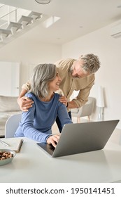 Happy Mature Senior 50s Couple Laughing Using Laptop Computer At Home. Smiling Middle Aged Older Husband Embracing Mid Age Wife Having Fun Doing Shopping Online, Working Or Browsing In Living Room.