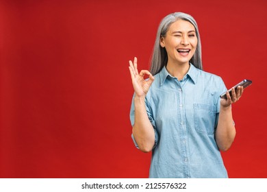 Happy Mature Senior Asian Woman Holding Smartphone Using Mobile Online Apps, Smiling Old Middle Aged Lady Texting Sms Message Chatting On Phone Isolated Over Red Background. Ok Sign.