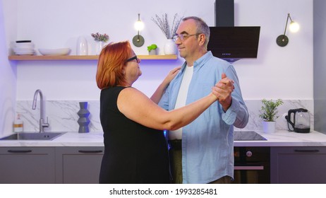 Happy Mature Senior Couple Dancing Laughing In The Kitchen, Beautiful Romantic Middle Aged Older Grandparents Relaxing Having Fun Together At Home Celebrating Anniversary Enjoy Care Love Tenderness.