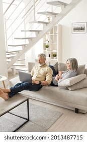 Happy Mature Senior Couple Using Devices Relaxing At Home On Sofa. Older Husband And Wife Using Devices Laptop And Phone Spending Free Time With Technology On Couch In Modern Apartment Living Room.