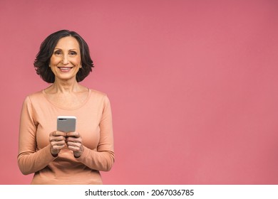 Happy mature senior woman holding smartphone using mobile online apps, smiling old middle aged lady texting sms message chatting on phone looking at cellphone isolated over pink background. - Powered by Shutterstock