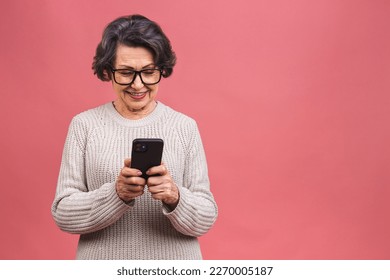 Happy mature senior woman holding smartphone using mobile online apps, smiling old middle aged grandmother texting sms message chatting on phone isolated over pink background.