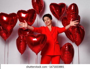 Happy Mature Short Hair Woman In Red Pantsuit Is Standing With Her Arms Spread Going To Hug Happy To Welcome Surrounded By Red Heart Air Balloons. Holiday Party.