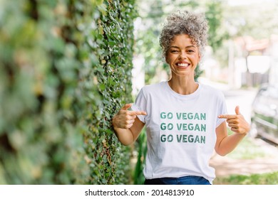 Happy mature vegan activist smiling cheerfully while standing alone outdoors. Cheerful mature woman advocating for veganism while wearing a shirt with the words "GO VEGAN" written on it.