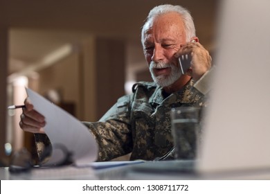 Happy Mature Veteran In Army Uniform Communicating On Cell Phone While Doing Paperwork In The Office.