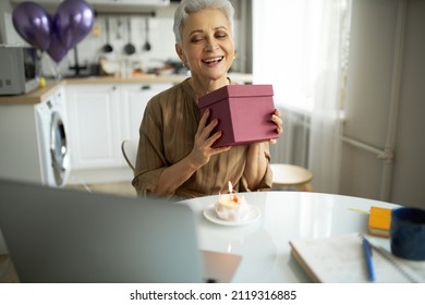 Happy mature woman of 50s smiling shaking present box to guess what's inside sitting in front of laptop and cupcake with candle against balloons in her kitchen, celebrating birthday online - Powered by Shutterstock