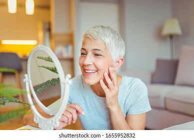 Happy Mature Woman Admiring Herself In The Mirror. Middle Aged Woman Looking At Wrinkles In Mirror. Plastic Surgery And Collagen Injections. Makeup. Good Skincare Habits Will Have You Looking Younger