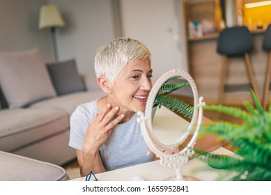 Happy Mature Woman Admiring Herself In The Mirror. Middle Aged Woman Looking At Wrinkles In Mirror. Plastic Surgery And Collagen Injections. Makeup. Good Skincare Habits Will Have You Looking Younger