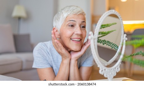 Happy Mature Woman Admiring Herself In The Mirror. Middle Aged Woman Looking At Wrinkles In Mirror. Plastic Surgery And Collagen Injections. Makeup. Good Skincare Habits Will Have You Looking Younger