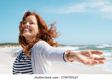 Happy mature woman with arms outstretched feeling the breeze at beach. Beautiful middle aged woman with arms up dancing on beach. Mid lady feeling good and enjoying freedom at sea, copy space.
