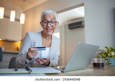 Happy Mature Woman Banking From Her Home Office. Old Woman Happy Doing Her Shopping Online Using A Credit Card. Portrait Of Beautiful Older Woman With Laptop Computer And Credit Card