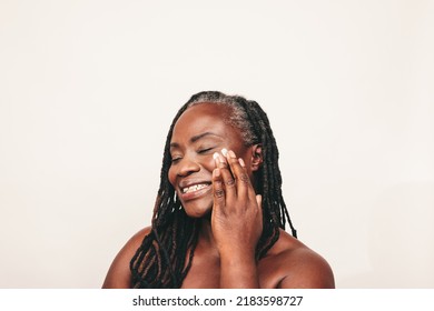 Happy Mature Woman With Dreadlocks Applying Moisturizing Cream On Her Face. Smiling Dark-skinned Woman Taking Care Of Her Flawless Melanated Skin. Cheerful Black Woman Ageing Gracefully.