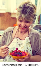 Happy Mature Woman Eating Berry Fruit Salad In The Kitchen.