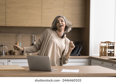 Happy mature woman enjoy track, dancing, stand at kitchen table with laptop, distracted from work, singing song, looking carefree alone at home. Modern tech, user of streaming digital music services - Powered by Shutterstock