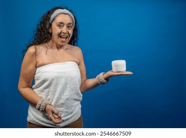 Happy mature woman with eye patches and towel wrapped around body, showing moisturizer cream jar after beauty treatment, on blue background - Powered by Shutterstock