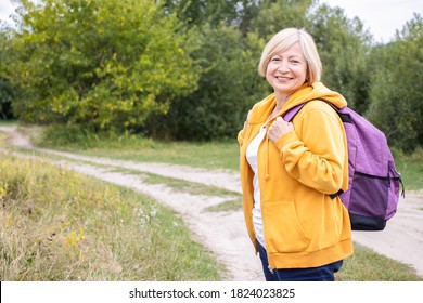 Happy Mature Woman Hiker With Backpack Behind Her Back, Wearing Yellow Hoodie, Smiling And Looking At Camera, Standing In Green Park, Forest Countryside Outdoors. Enjoying Active Travel Trip