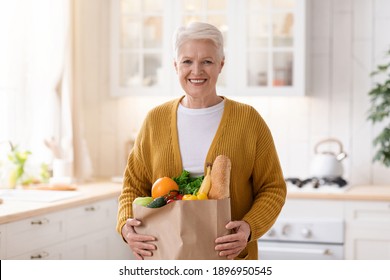 Happy Mature Woman Holding A Paper Bag Full Of Groceries From The Supermarket, Kitchen Interior. Elderly Woman With Groceries At Home, Food Delivery During Quarantine Concept, Copy Space