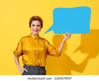 Happy Mature Woman Holds Blue Speech Bubble Banner. Photo Of Kind Attractive Woman In Yellow Shirt On Yellow Background