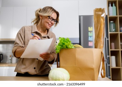 Happy Mature Woman In Kitchen Holding Shopping List Looking Through Items