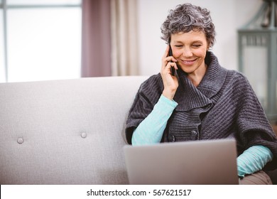 Happy Mature Woman With Laptop Using Mobile Phone While Sitting On Sofa At Home. Working From Home In Quarantine Lockdown. Social Distancing Self Isolation