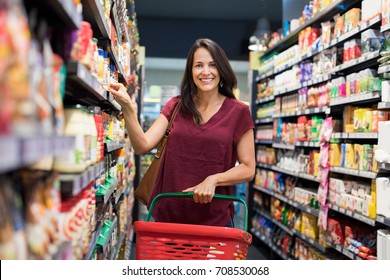 Happy mature woman looking at camera while shopping in grocery store. Casual woman choosing food from shelf in supermarket and looking at camera. Smiling customer standing near shelves.
