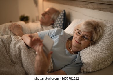 Happy Mature Woman Lying Down In Bed And Using Mobile Phone. The Man Is Sleeping In The Background. 