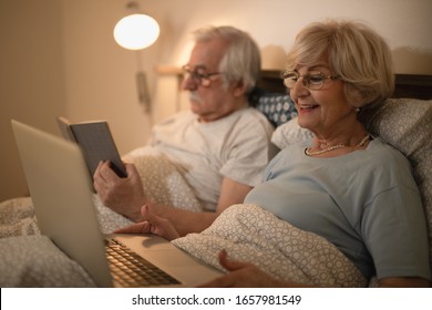 Happy Mature Woman Lying Down In Bed And Using Laptop At Night. Her Husband Is Reading A Book. 