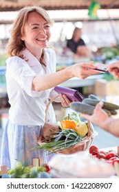 Happy Mature Woman Paying For Fresh Organic Vegetables In A Local Marketplace. Carrying Basket Filled With Vegetables. Healthy Organic Local Food Production