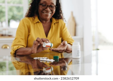 Happy mature woman sitting at her kitchen table and taking daily medication to manage a chronic condition. Woman adhering to her prescribed treatment plan to maintain her health and quality of life. - Powered by Shutterstock