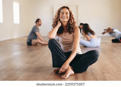 Happy mature woman sitting in a yoga studio with a group of people working out in the background. Smiling senior woman having an exercise session with her class in a community yoga studio. - Powered by Shutterstock