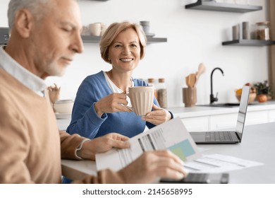 Happy Mature Woman Smiling, Drink Coffee, Sitting Near Her Husband While He Paying Utility Bills. Concept Of Social Benefits, Financial Security And Pension Reform. Married Family Couple Checking Tax