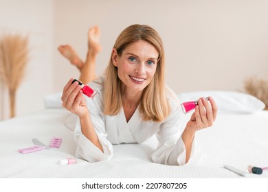 Happy Mature Woman In White Robe Lying On Bed And Choosing Nail Polish, Making Manicure In Bedroom Interior, Copy Space. Cosmetics, Beauty Care At Home