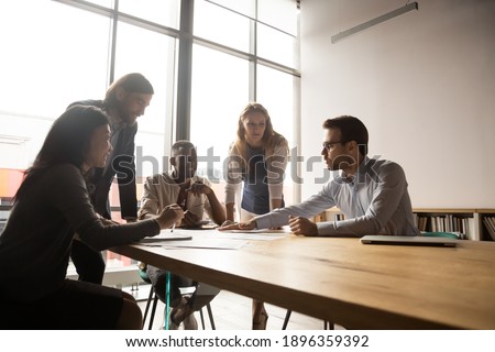 Happy mature and young multiracial employees listening to skilled 30s male team leader, developing corporate strategy together, brainstorming project ideas or analyzing research results in office.